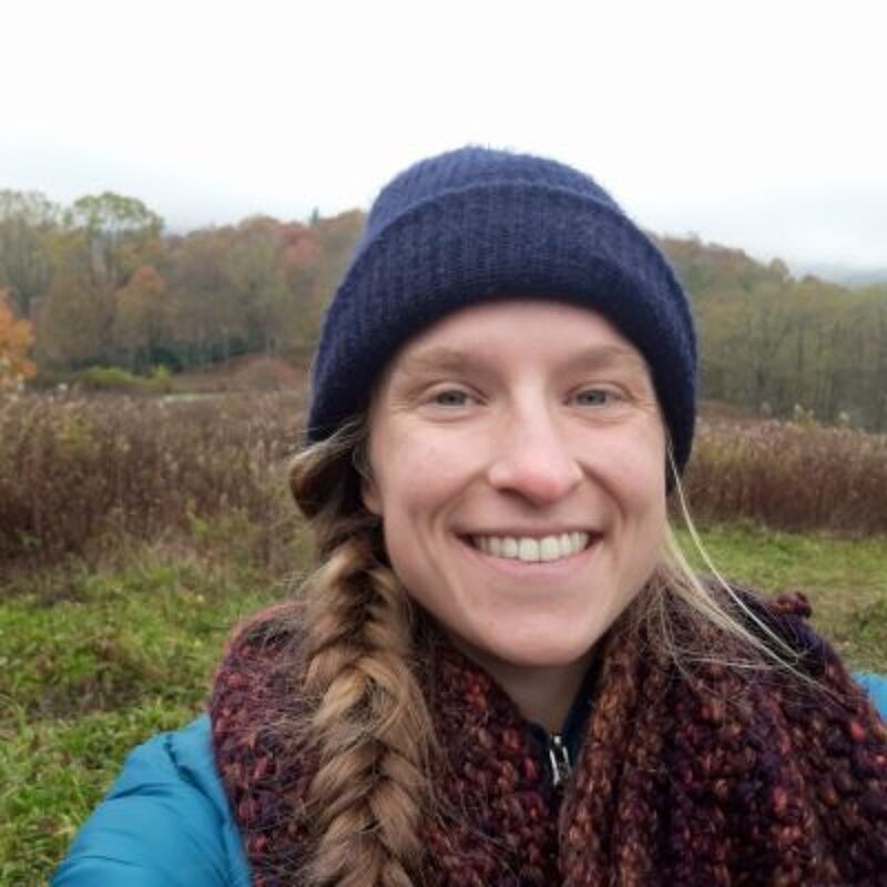 The image shows a woman with blonde hair braided to the side, wearing a blue beanie and a colorful scarf. She is smiling at the camera. The background features a field with tall grasses and trees with autumn foliage, suggesting it's taken during the fall season. The sky appears overcast.
